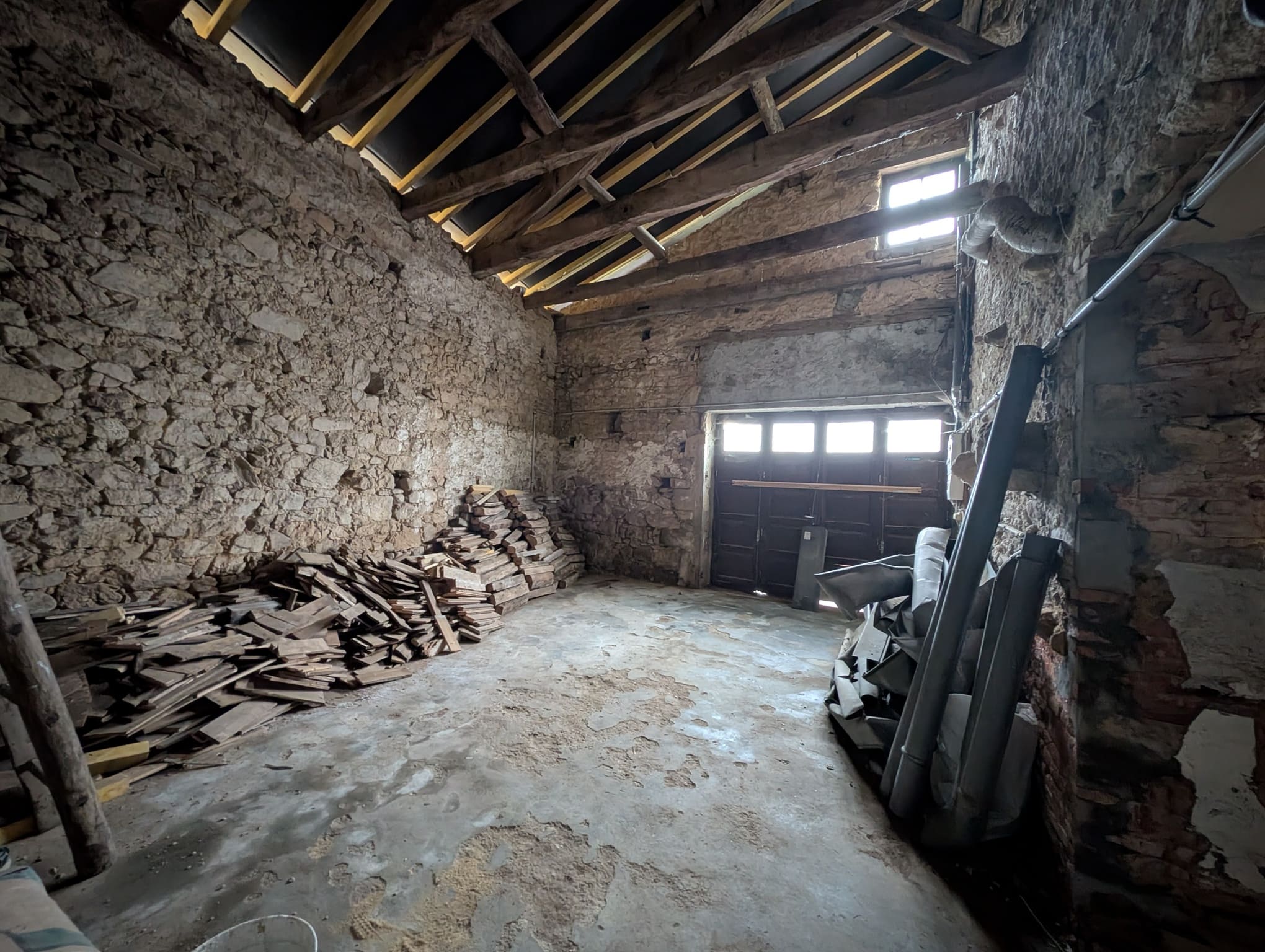 Intérieur du garage avant transformation, murs en pierre, charpente bois visible, porte de garage bois existante.