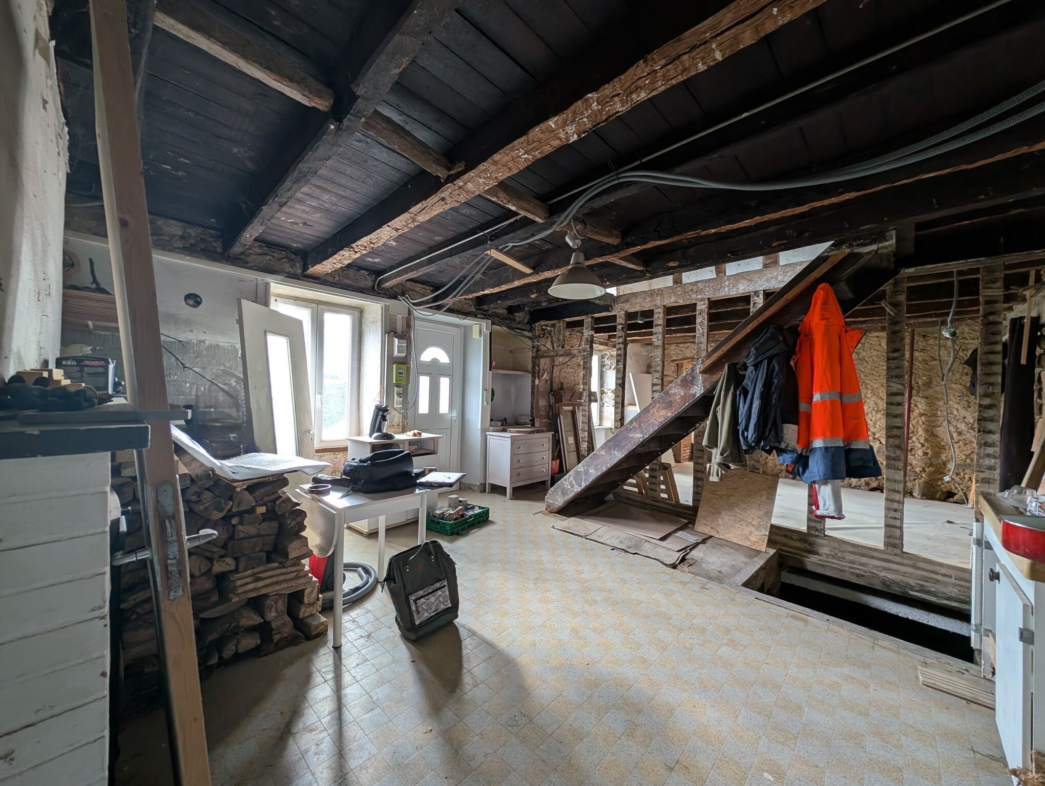 Vue de la pièce principale au rez-de-chaussée après démolition, poutres apparentes et escalier en bois ancien.