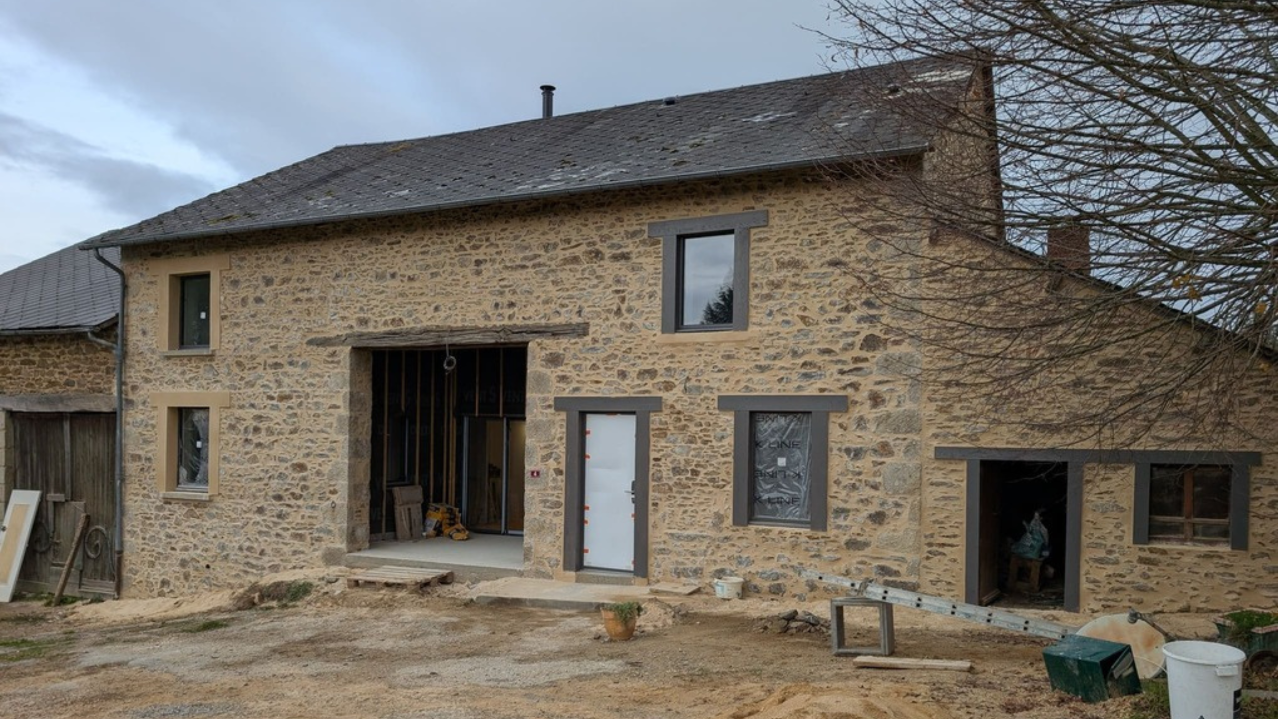 Vue extérieure de la grange en pierre pendant le chantier, avec les ouvertures recréées, les nouvelles menuiseries posée et le rejointoiement révélant l’évolution du bâti agricole vers une habitation.