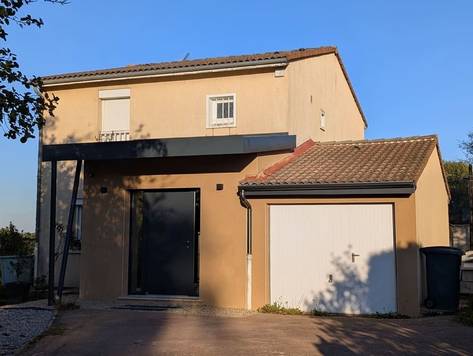 Façade principale rénovée avec nouvelle extension d’entrée en toiture plate, porte d’entrée gris anthracite et auvent métallique contrastant avec la toiture tuilée du garage accolé.