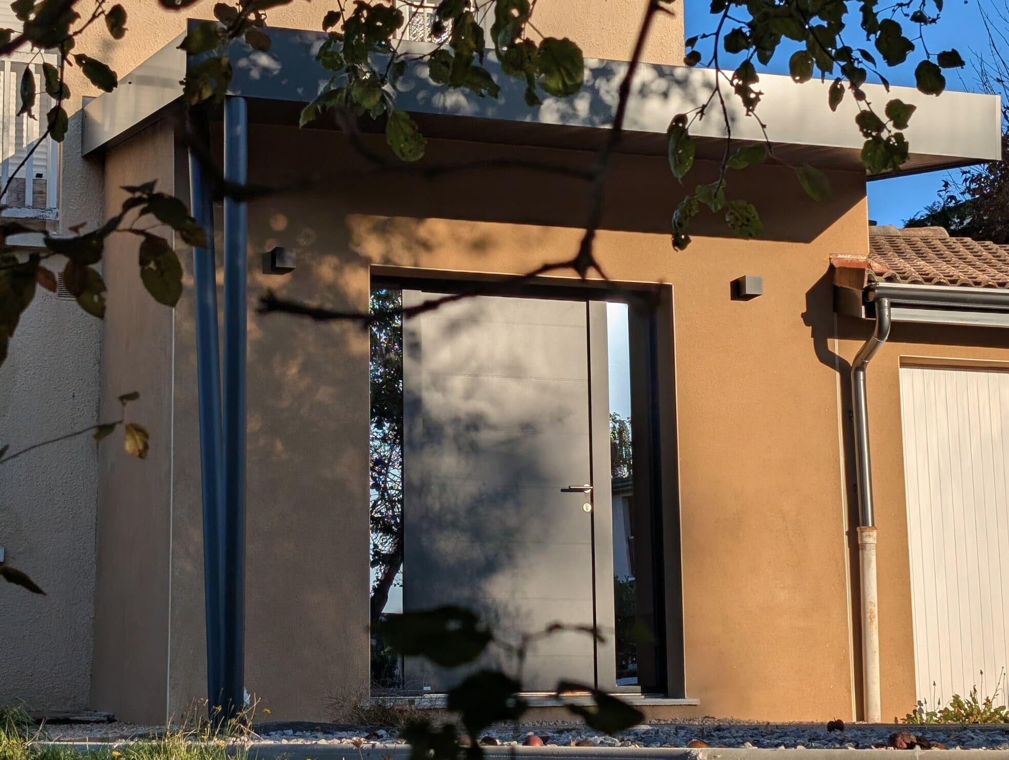 Vue extérieure de l’extension contemporaine créée pour agrandir l’entrée de la maison, avec casquette métallique, enduit chaleureux et porte d’entrée anthracite.